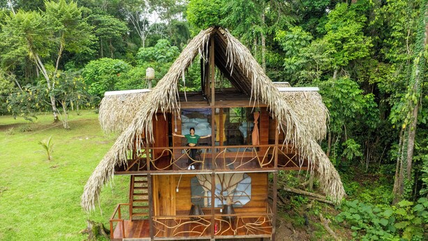 Une maison en bois à deux étages, dotée d’un toit de chaume et entourée d’une végétation luxuriante.