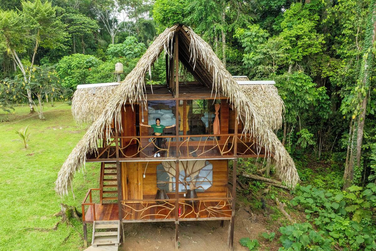 A two-story wooden house with a thatched roof, surrounded by lush greenery.