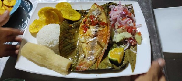 Une assiette contenant un poisson enveloppé dans des feuilles de bananier, accompagné de riz, de bananes plantains en tranches et d’une garniture d’oignons et de tomates coupés en dés.