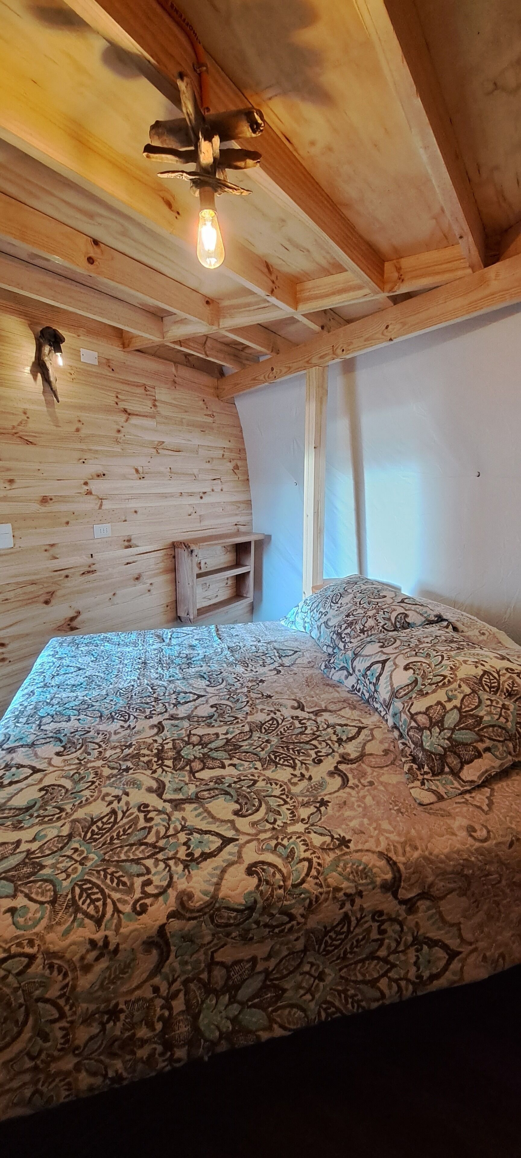 Una habitación de cabaña de madera con una cama, un pequeño estante de madera y una lámpara.