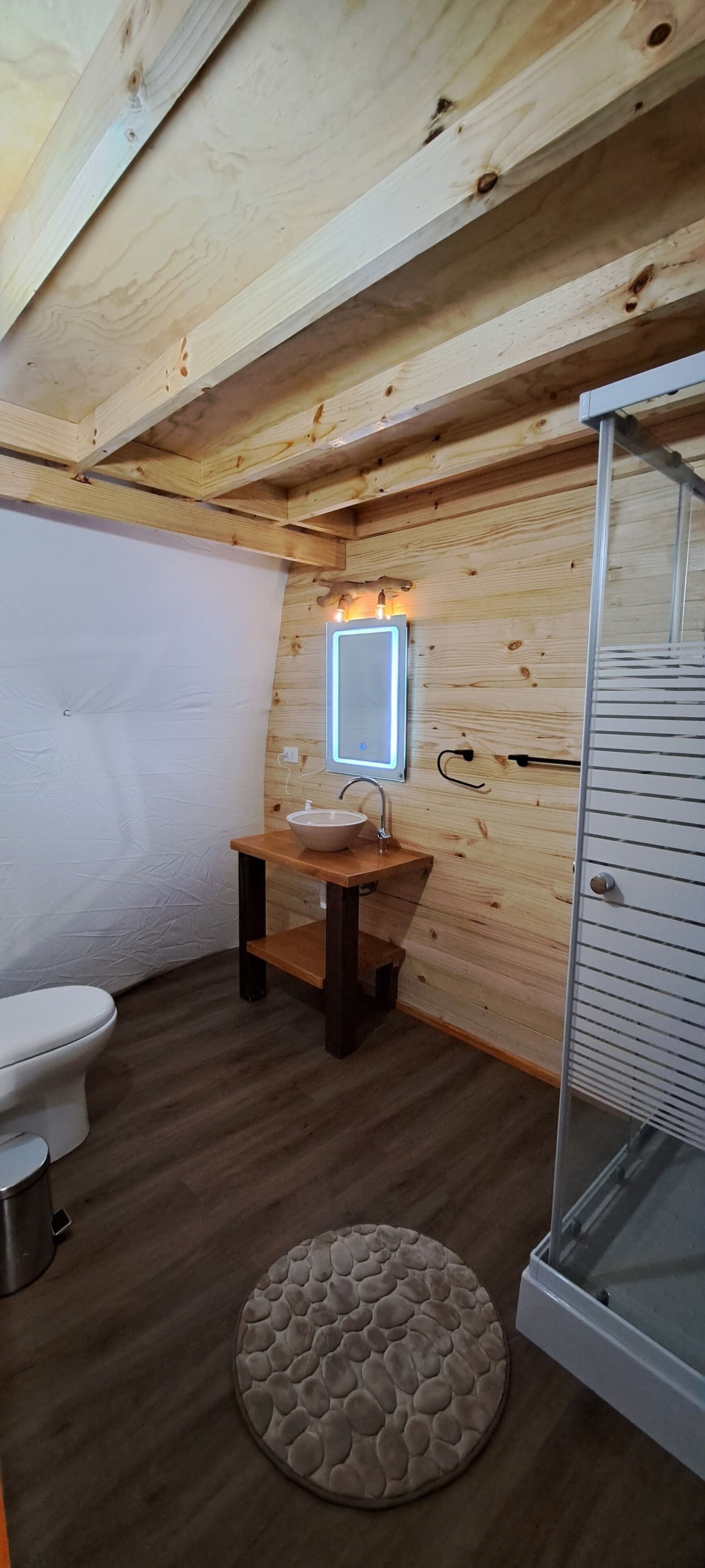 Ein kleines, rustikales Badezimmer mit Holzbalkendecke, einem weißen Waschbecken, einer Toilette und einer Duschkabine aus Glas.