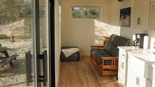 A compact kitchen with wooden cabinets, a built-in bench, and a window overlooking a sandy outdoor area. - Trout Container Home - Fish Camp Rockport (Rockport)