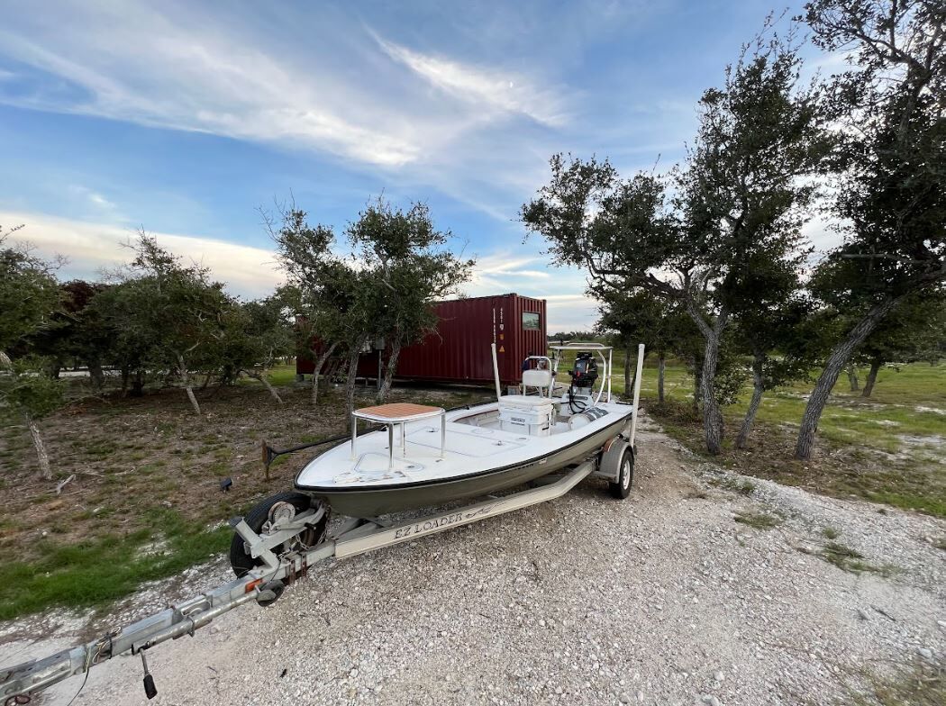 A boat trailer with a boat on it, parked on a gravel path with trees in the background.