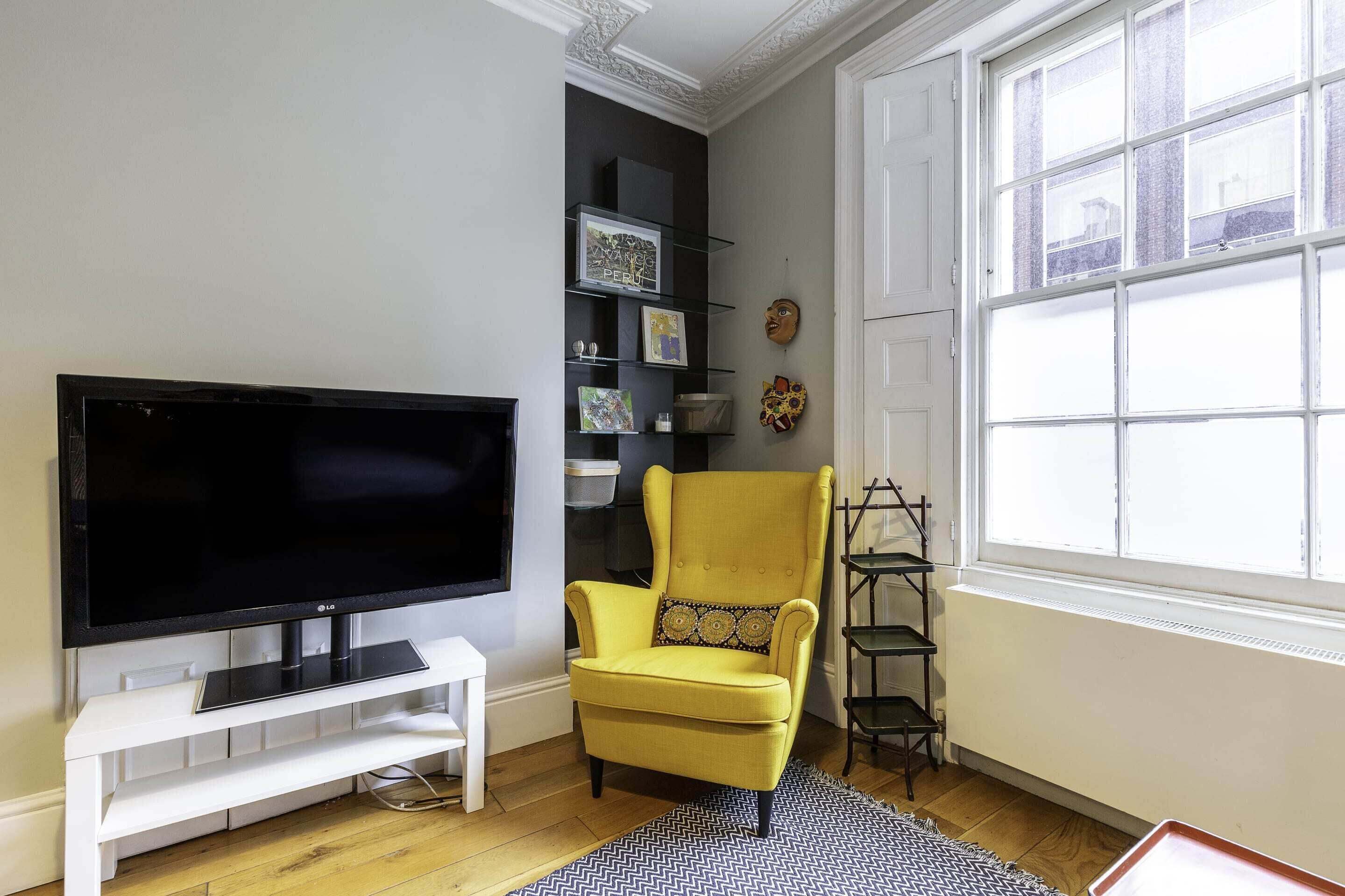 A living room with a flat-screen TV on a white stand, a yellow armchair with a patterned cushion, and a black and white rug.