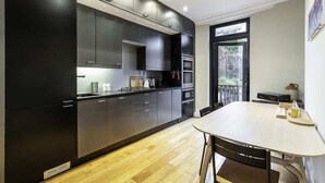 A modern kitchen with dark cabinets, a wooden dining table, and a view of the outdoors through a large window. - Stylish Kings Cross Apartment With Garden Retreat (London)