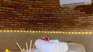 Una habitación con pared de ladrillo, una cama con ropa de cama blanca, una mesita con velas y aceites esenciales, y un equipo de aire acondicionado.