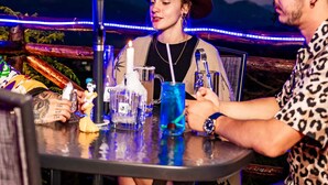 Dos personas sentadas en una mesa al aire libre, con una bebida y una vela, bajo una sombrilla azul, con montañas al fondo.