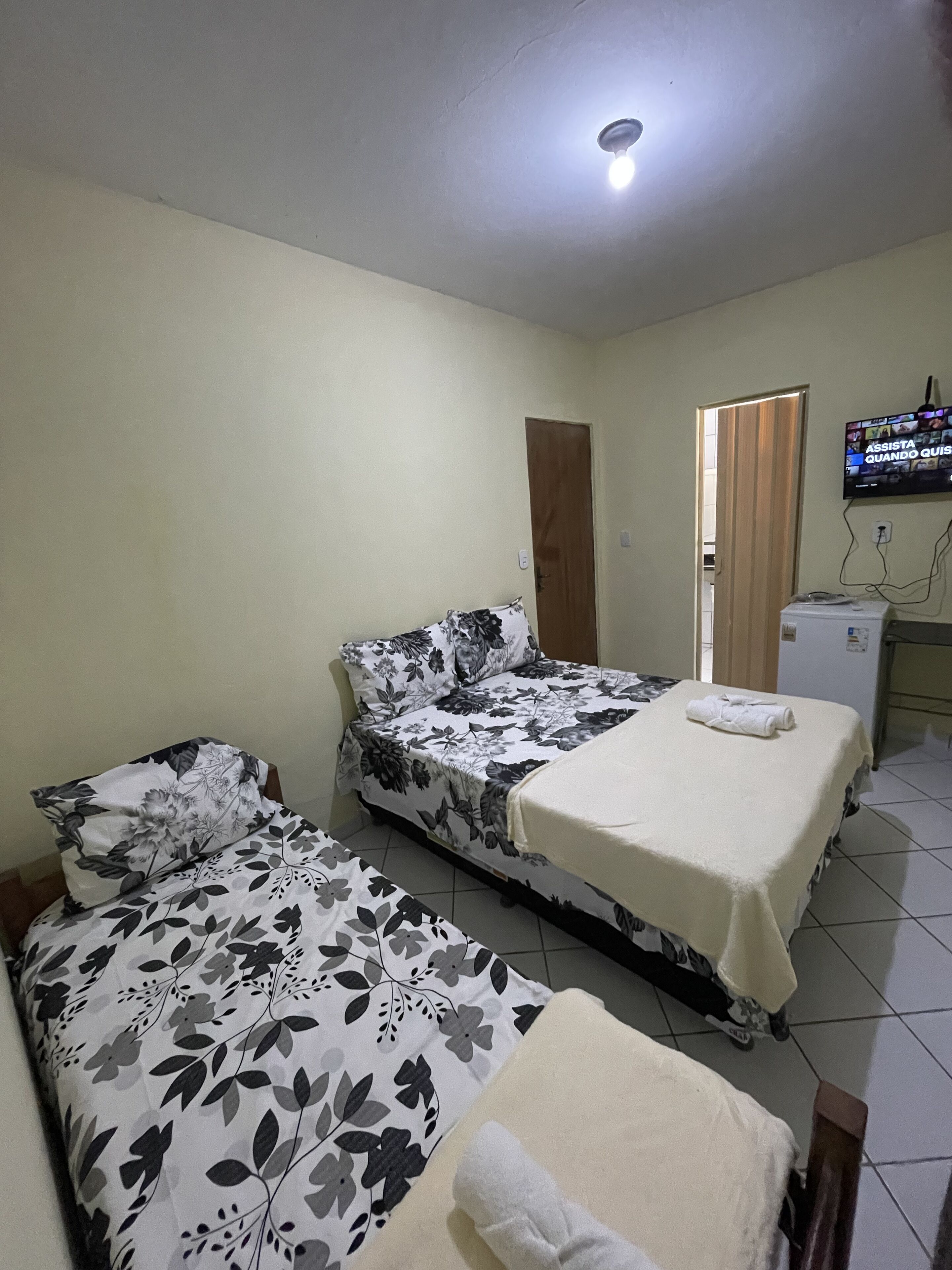 Quarto com duas camas, cada uma com colcha florida e manta branca. Há uma televisão na parede e um frigorífico pequeno no canto.