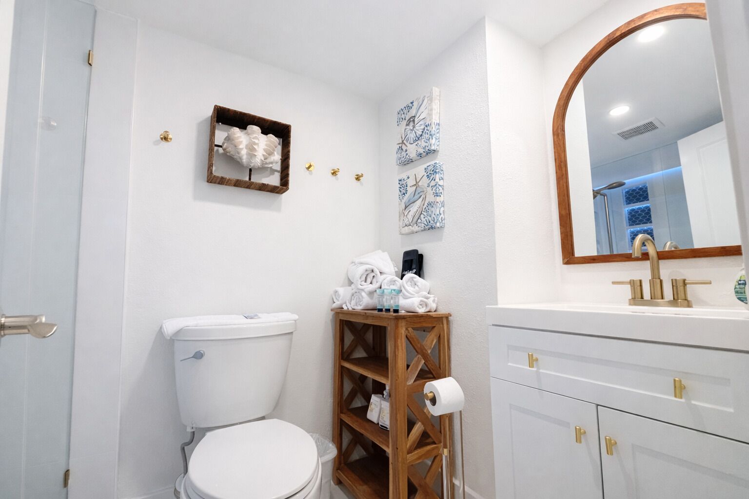 Une salle de bain comprenant des toilettes, une étagère en bois avec des serviettes, un miroir et un lavabo avec des robinets dorés.