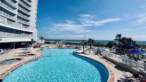 A large, clear swimming pool with a curved design, surrounded by lounge chairs and umbrellas, and a high-rise building with balconies in the background.