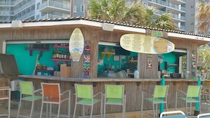 A beachside bar with a wooden facade, bar stools, and a menu board.