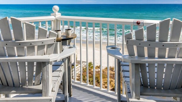 A deck with white Adirondack chairs, a small table with a cup and saucer, and a view of the ocean.