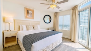 A hotel room with a large bed, a ceiling fan, a nightstand with a lamp, a window with curtains, and a view of the city.