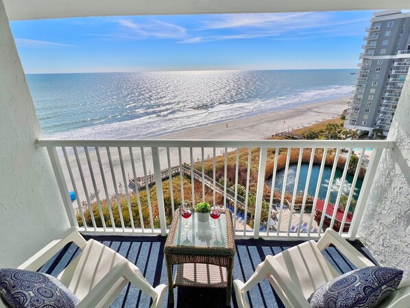 A balcony with a view of the ocean and a pool. There are two chairs and a table with a glass top.