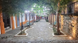 A cobblestone pathway flanked by trees and benches, leading to a building with a stone wall and a metal fence.