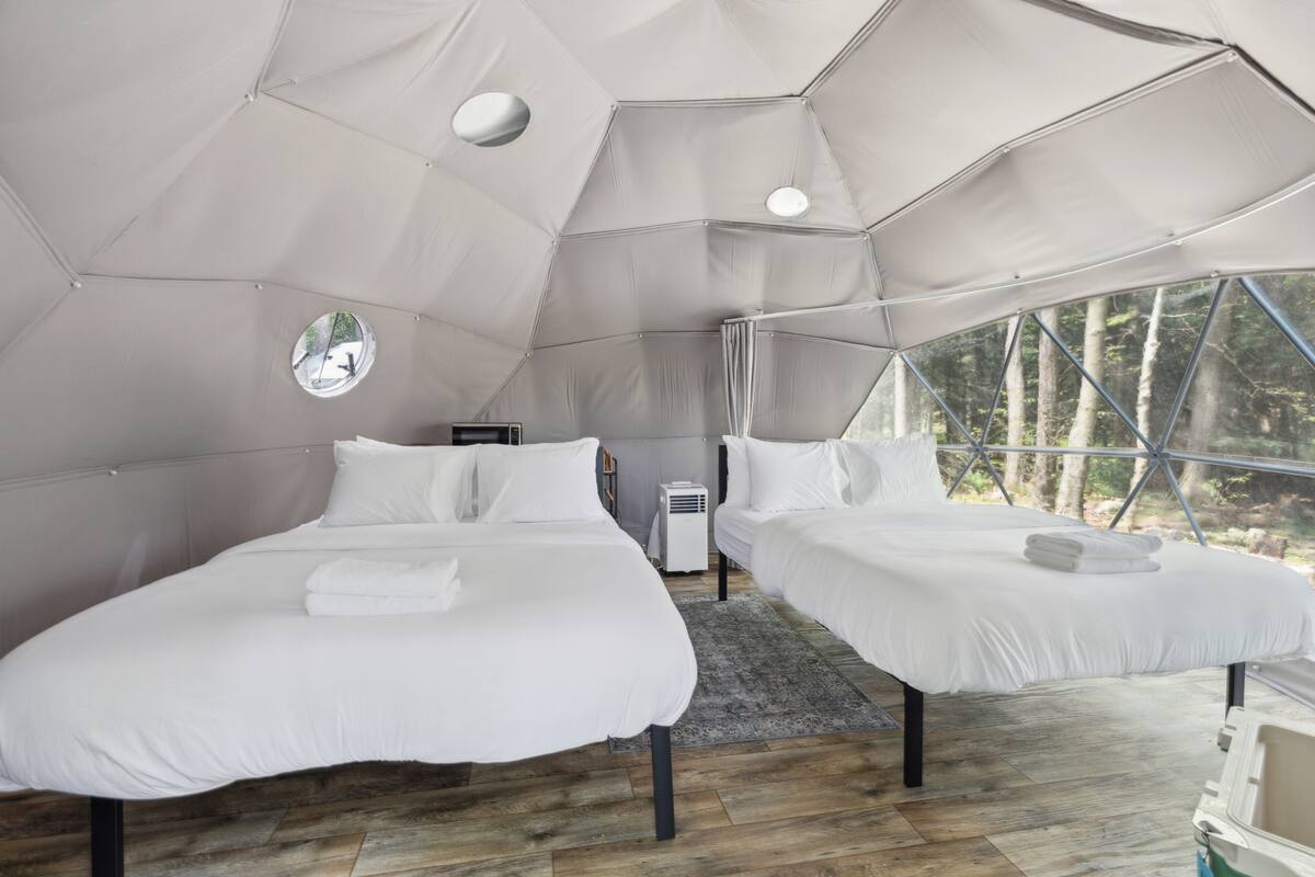 A tent-style room with two beds, a small circular window, and a view of trees outside.