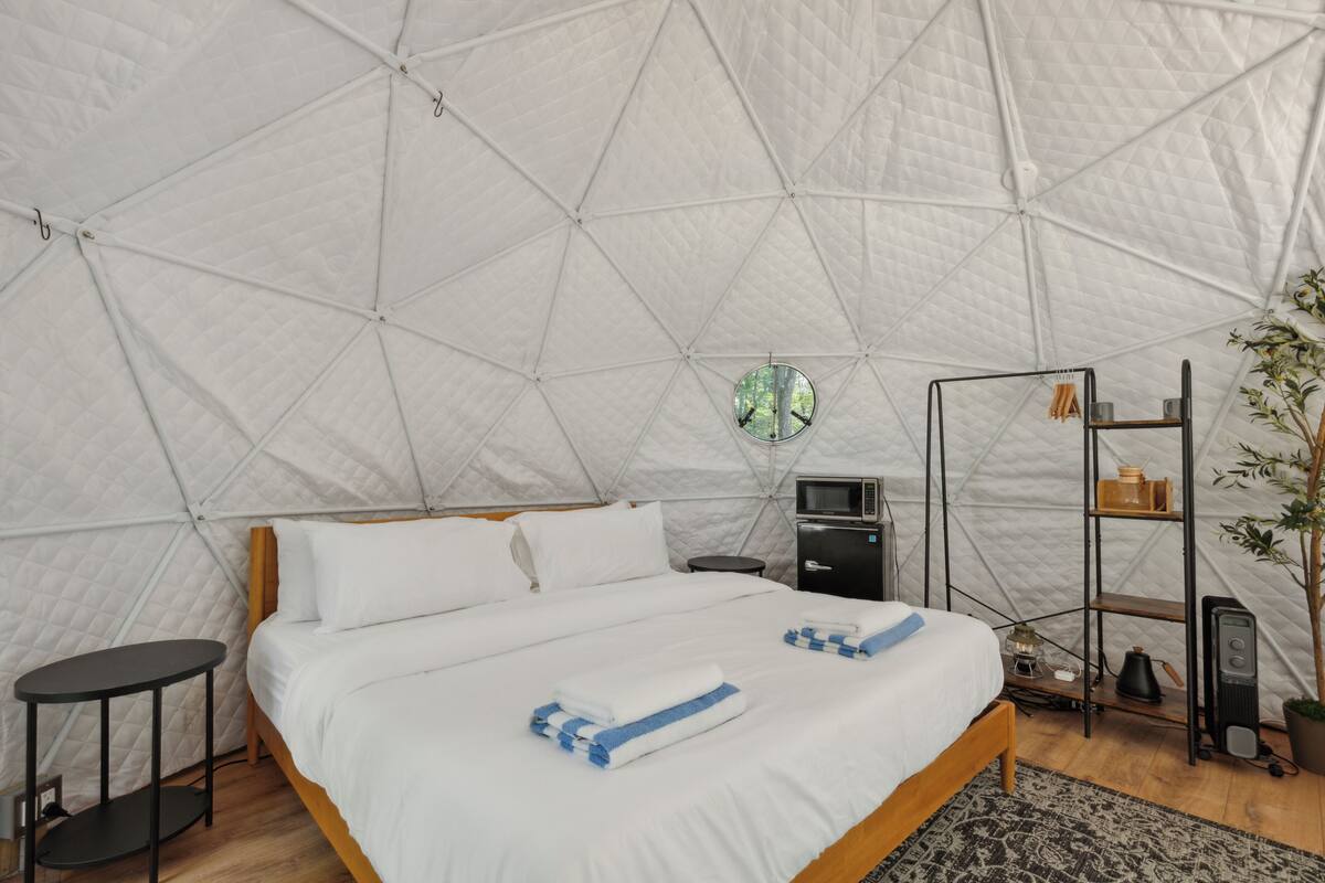 A tent-style room with a bed, a small table, a shelf with items, and a potted plant.