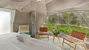 A tented room with a bed, two chairs, a small table with a plant, and a window with a forest view. - Ferncrest Campground (Greentown)