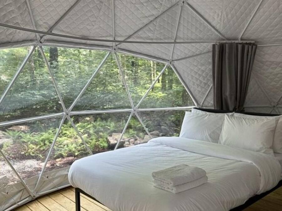 Ein Zimmer mit einem Bett, weißer Bettwäsche und Blick auf einen Wald durch ein großes Fenster.