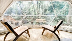 A geodesic dome structure with two wooden chairs facing a wooden table, set on a wooden floor with a view of a forested area through large windows. - Ferncrest Campground (Greentown)