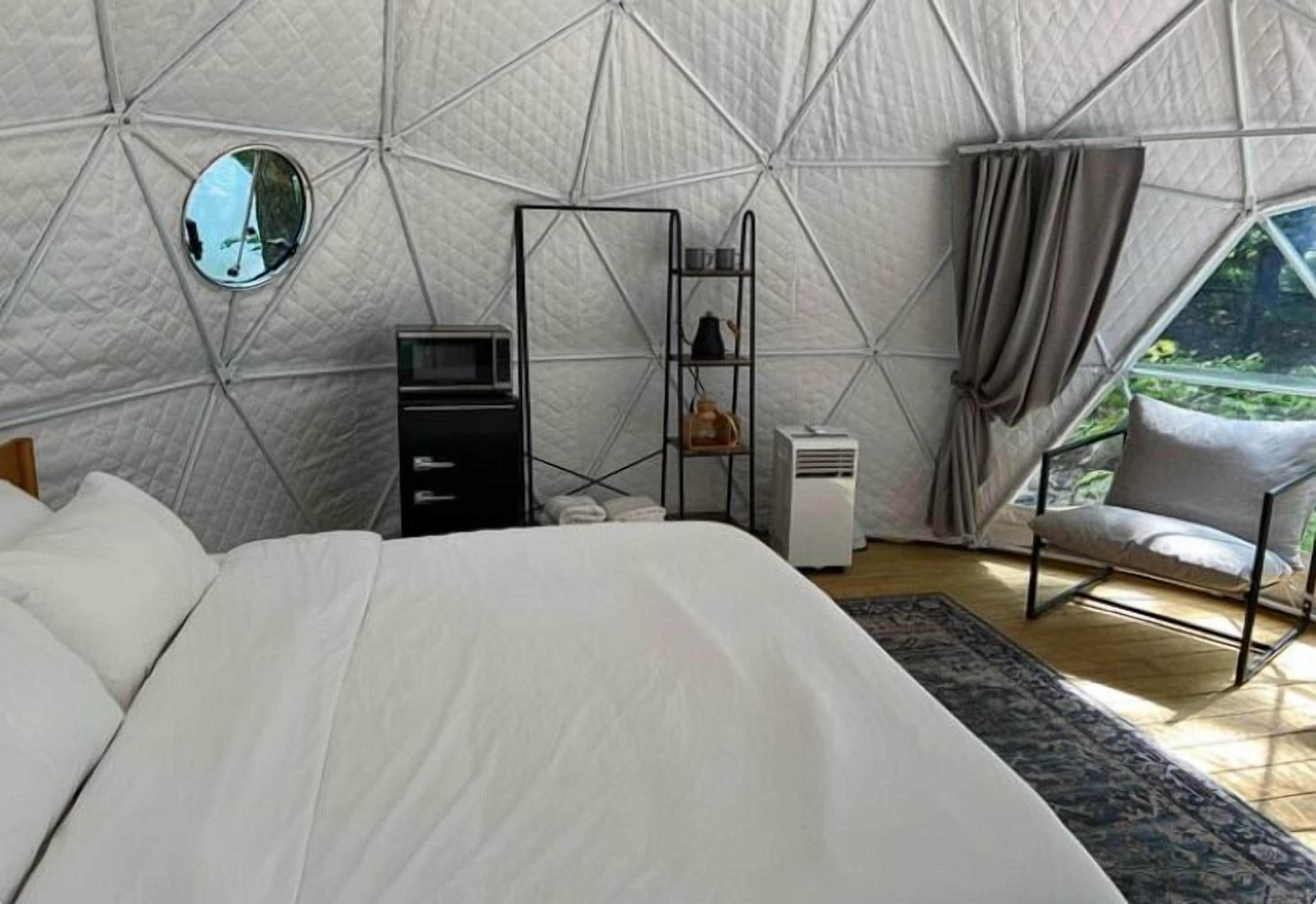 Un dormitorio con cama, televisor, estante con objetos, una silla y una ventana.