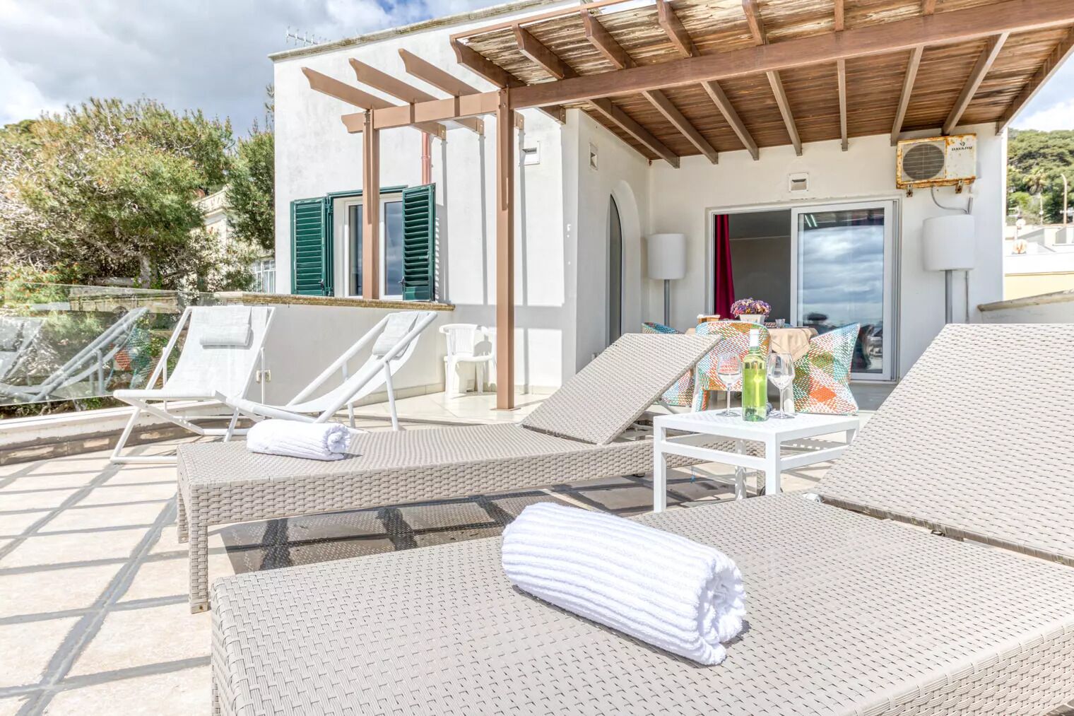 A rooftop terrace with lounge chairs, a table with a bottle, and a view of a building with shutters.