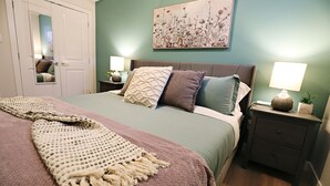 Un dormitorio con una cama, dos mesitas de noche, una mesita auxiliar, una puerta y un cuadro colgado en la pared.