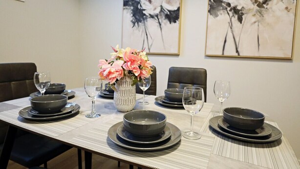 Una mesa de comedor dispuesta con platos negros, tazones y copas de vino, junto a un florero con flores.