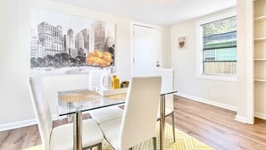 A dining area with a glass table, white chairs, a cityscape painting, and a window with blinds. - Private Birmingham Retreat Near Red Mountain Park!

 (Birmingham)