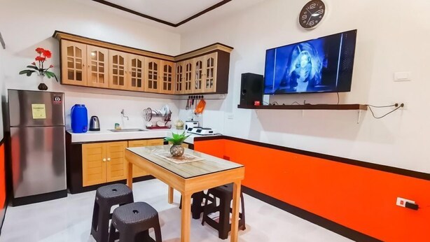 Cozinha compacta com armários de madeira, geladeira de aço inoxidável, mesa de jantar com banquetas e televisão na parede.