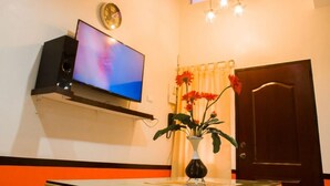 Quarto com televisão de tela plana na parede, porta de madeira, janela com cortinas e um vaso com flores sobre uma mesa.