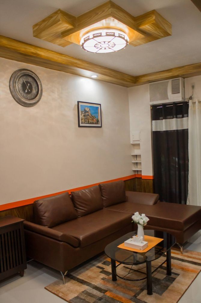 Un salon avec un canapé en cuir marron, une petite table sur laquelle est posé un vase, une horloge au mur et une fenêtre avec des rideaux.