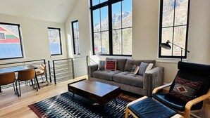 A spacious living room with a high ceiling, large windows offering mountain views, a ceiling fan, and a cozy seating area with a sofa and armchair. - New Luxury Home in the Heart of Ouray, Walk Main St, Breathtaking Views, AC (Ouray)
