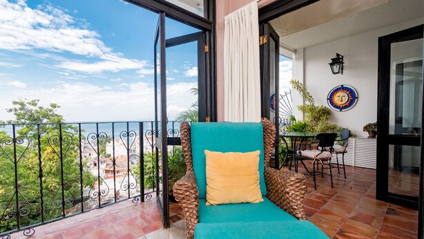 A balcony with a view of the city, a teal chair with a yellow pillow, and a small table with chairs.