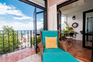 Ein Balkon mit Blick auf die Stadt, ein türkisner Stuhl mit gelbem Kissen und ein kleiner Tisch mit Sitzgelegenheiten.