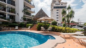 A swimming pool with lounge chairs and a thatched-roof pavilion, surrounded by a landscaped garden and adjacent multi-story buildings.