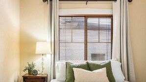 A hotel room with a bed, a nightstand with a lamp, a window with blinds, and a view of the outside. - Quiet Home Near Downtown + Mountain Views (Salt Lake City)