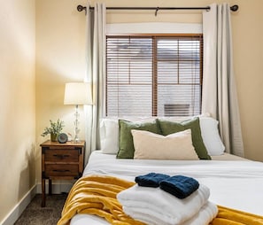 Une chambre d’hôtel avec un lit, une table de chevet avec une lampe, une fenêtre avec des stores et une vue sur l’extérieur.