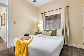 A hotel room with a bed, a nightstand, a lamp, a window with blinds, and a door leading to another room.
