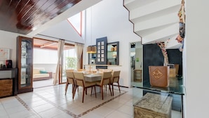 Un comedor amplio con techo alto y vistas al exterior. Hay una mesa de comedor con sillas, un aparador de vidrio con objetos decorativos y un espejo grande en la pared.
