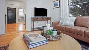 A living room with a round coffee table, a television on a stand, a brown sofa, and a window with a view of trees. - Whitetail Bungalow | Walk to Downtown Nampa | King Bed (Nampa)