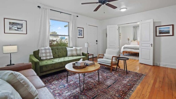 A well-lit room with a green sofa, a white armchair, a wooden coffee table, and a ceiling fan. - Whitetail Bungalow | Walk to Downtown Nampa | King Bed (Nampa)