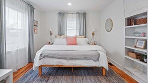 A bedroom with a bed, bedside tables, a nightstand, a bookshelf, and a window with curtains. - Whitetail Bungalow | Walk to Downtown Nampa | King Bed (Nampa)