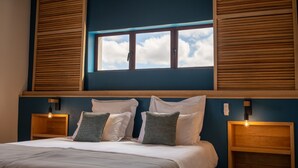 Un dormitorio con una cama, dos mesitas de noche, un ventanal con persianas y vista al cielo.
