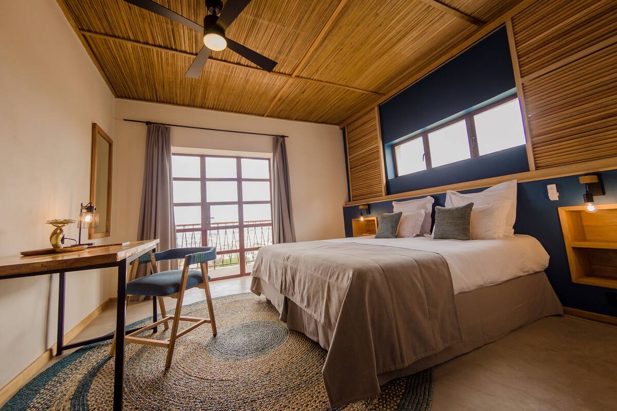 A bedroom with a bed, desk, chair, and a large window.