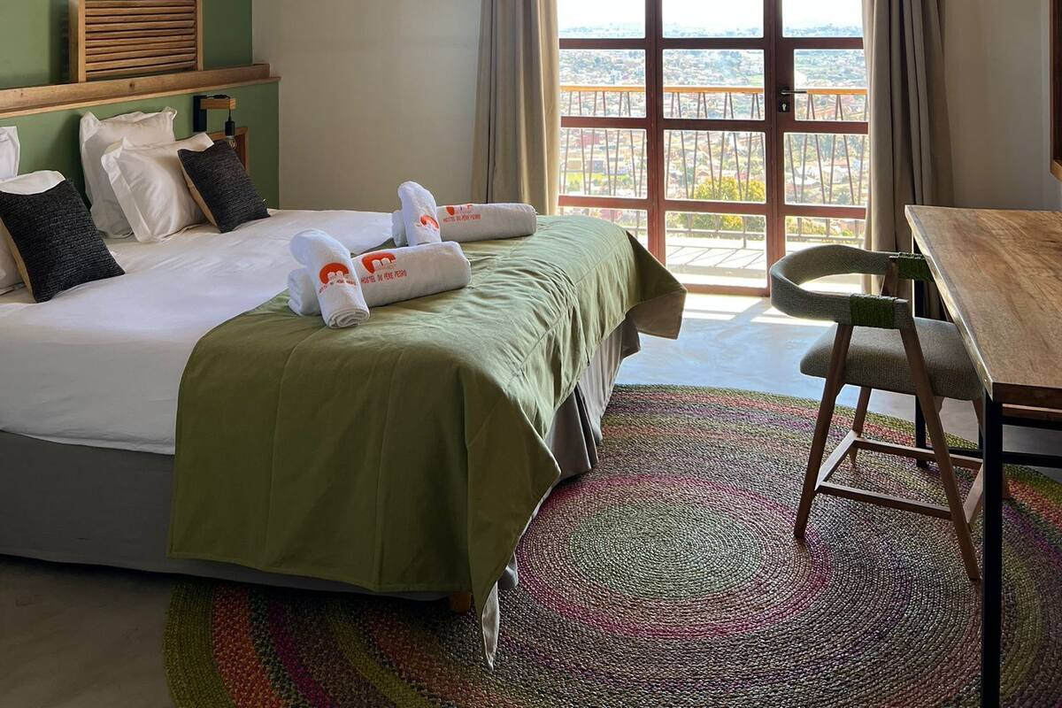 A hotel room with a bed, a table, a chair, a ceiling fan, and a large window with curtains.