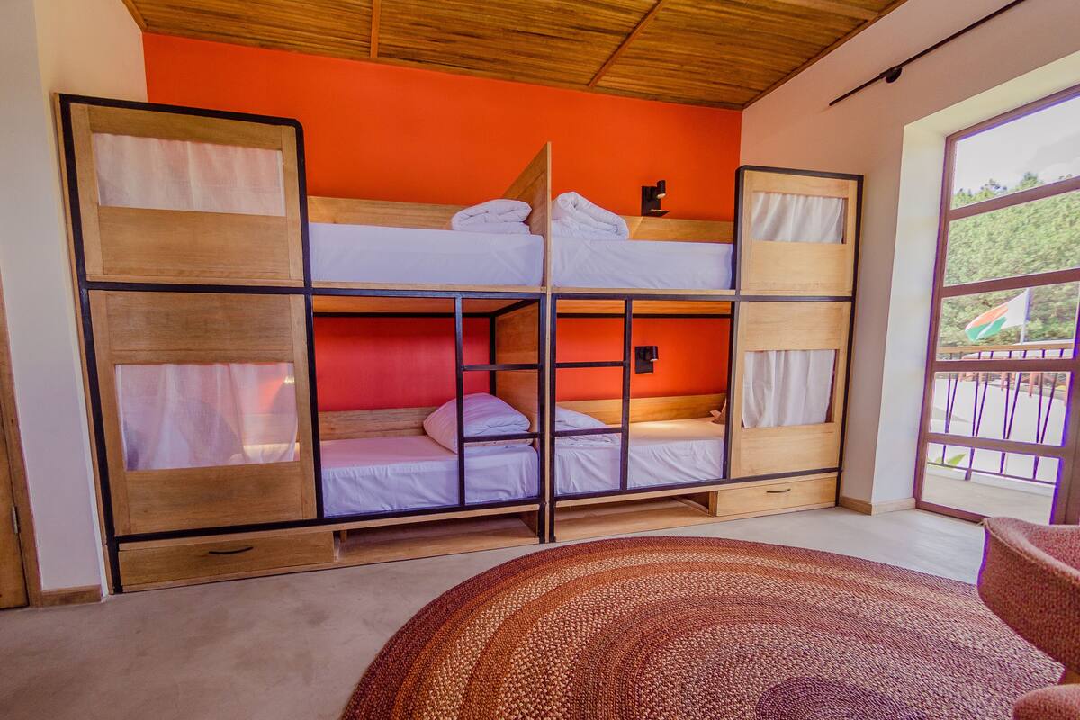 A bunk bed room with a wooden frame, a red accent wall, and a large window with a view of greenery.