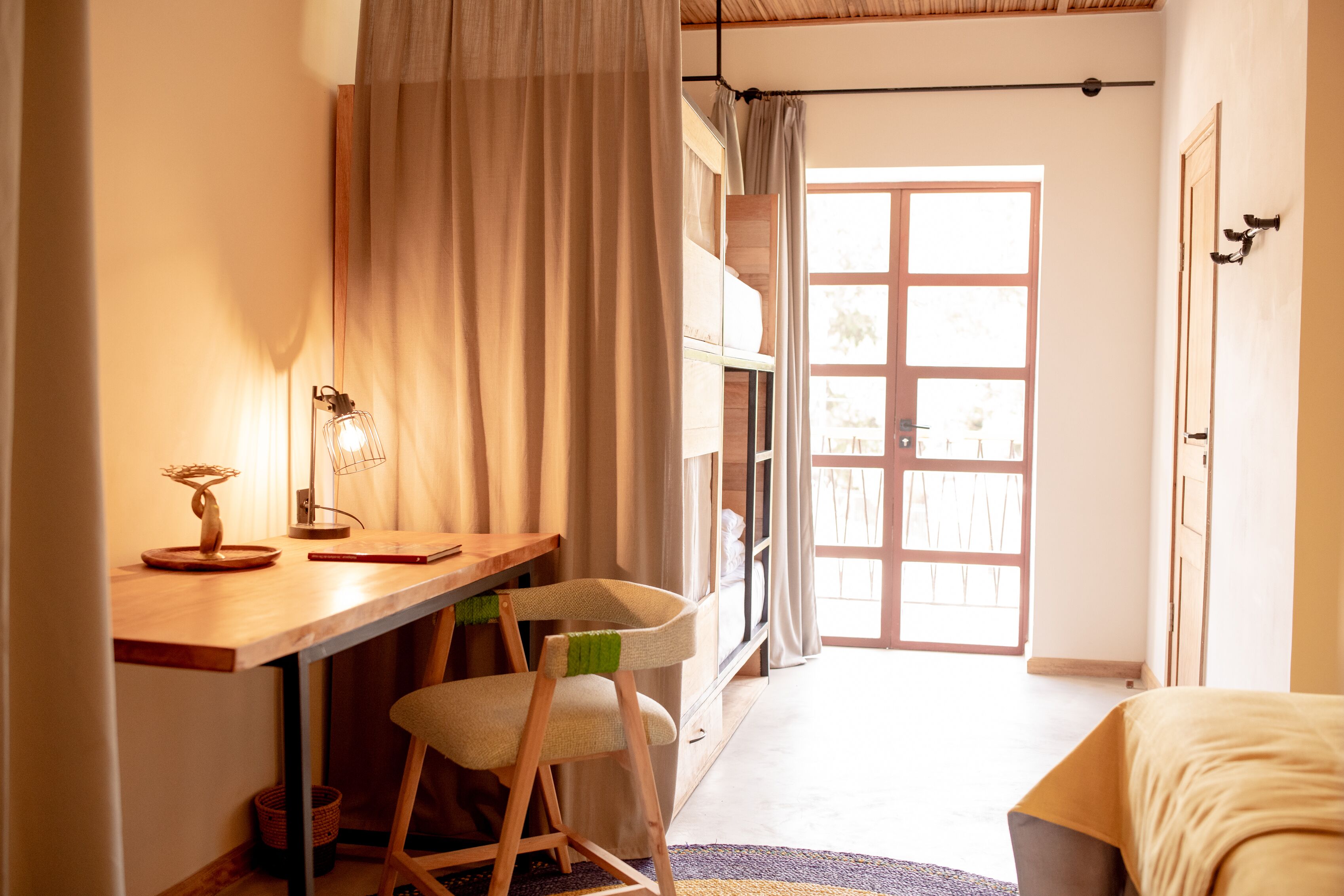Une pièce avec un bureau en bois, une chaise, un lit et une grande fenêtre avec des rideaux.
