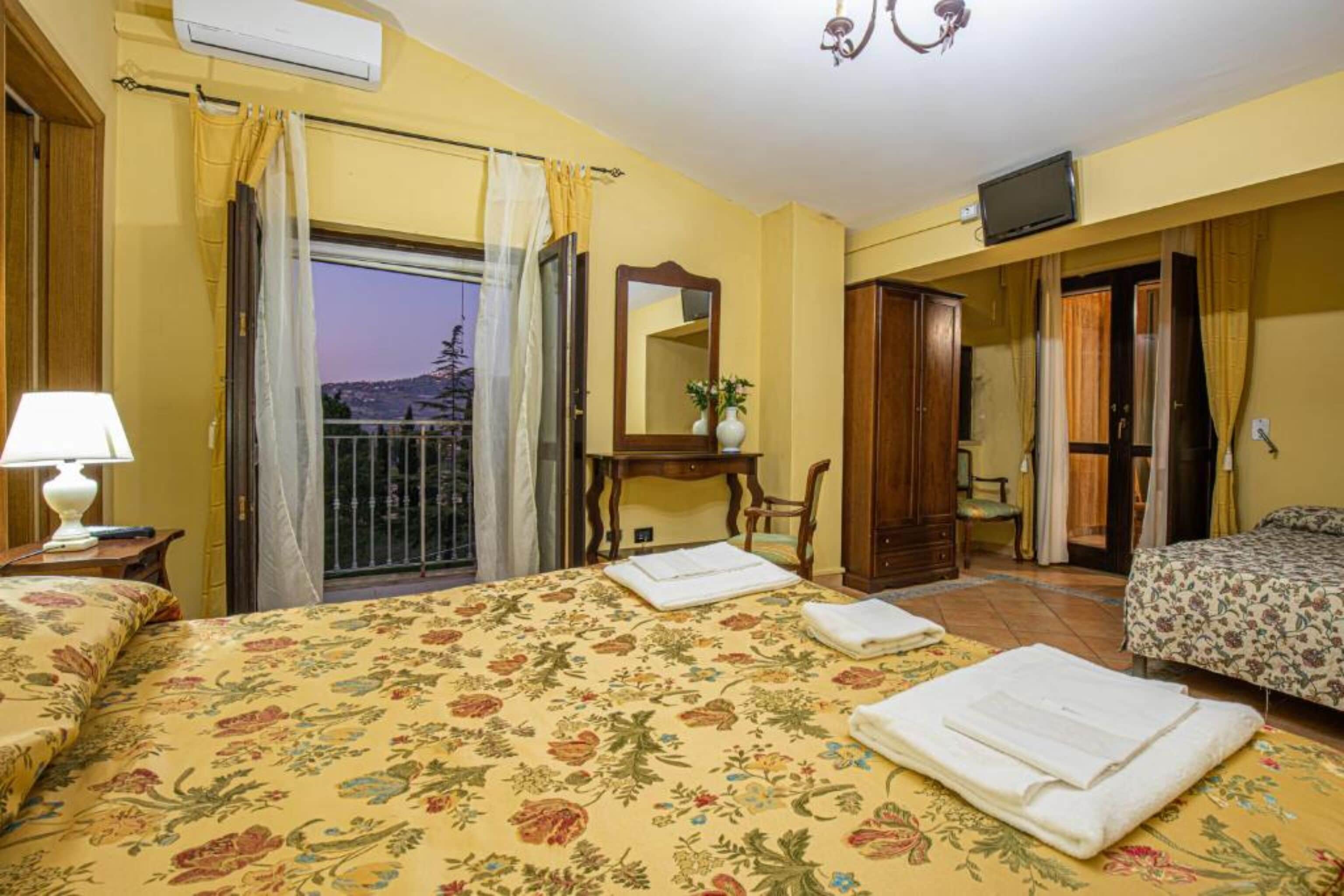 Una habitación de hotel con dos camas, un escritorio, una silla, televisión y un balcón con cortinas.