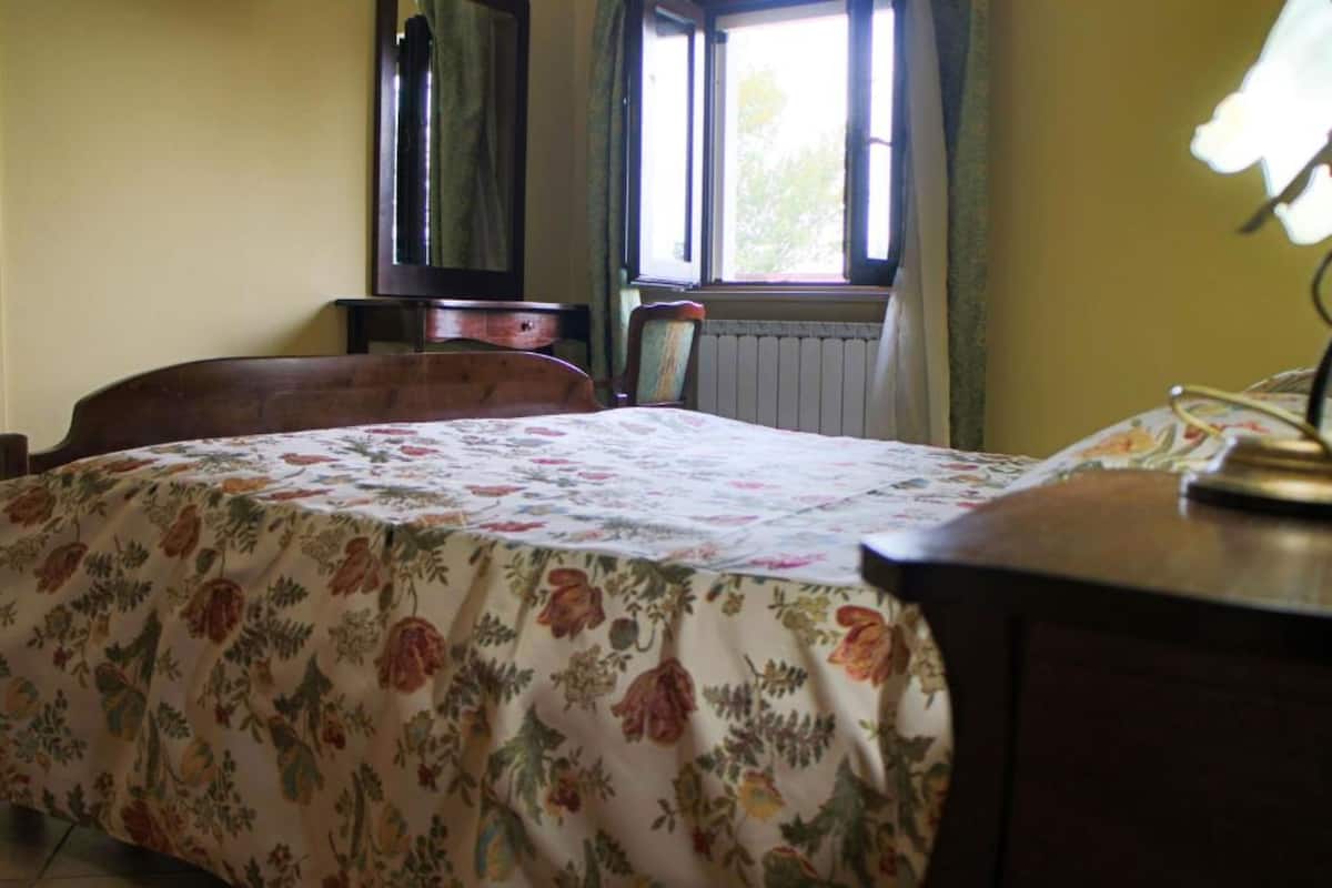 A bedroom with a floral bedspread, a wooden headboard, a nightstand, a chair, and a window with curtains.